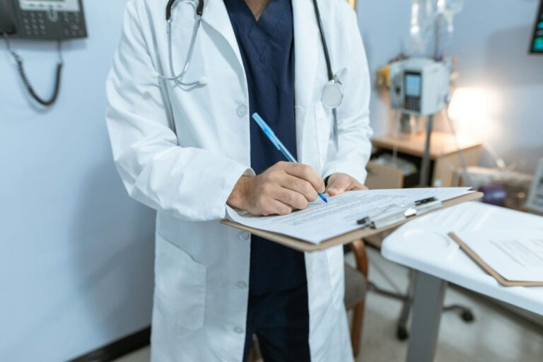 A doctor in a lab coat writing notes in a hospital setting, using a clipboard.